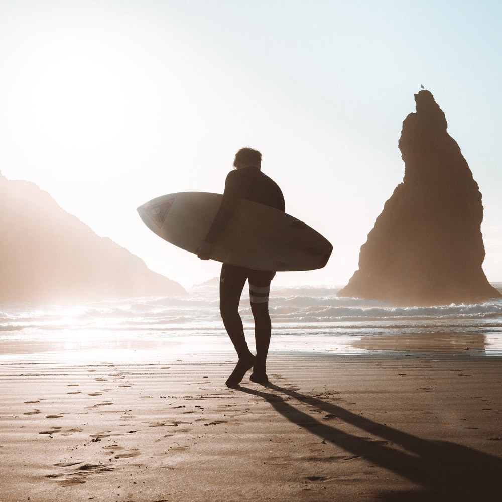 Image of surfer walking on beach silhouetted by the sun.